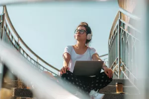 Female freelancer wearing headphones working on the laptop and drinking coffee. Sunny day in the city. Generation Z concept.generacija Z, gen z, mladi, mladež, nove generacije, mladost, mlađi