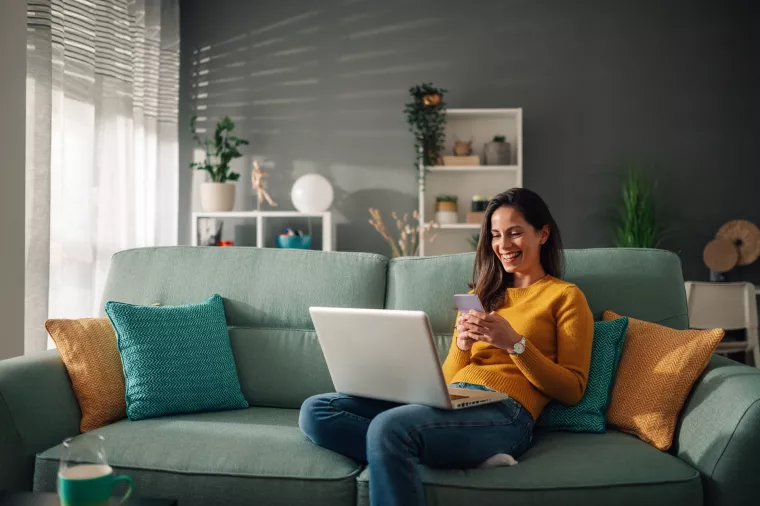 Smiling caucasian woman using laptop and typing on a phone while sitting on couch at home. Happy female shopping or chatting online in social network, typing blog, sending message or email. Copy space