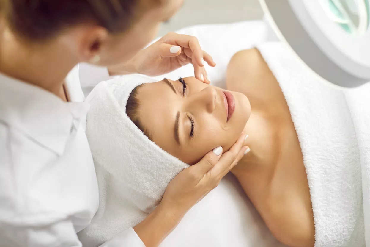 Young professional woman cosmetologist doing skin care facial treatment examining its condition for female client with special lamp in beauty clinic. Cosmetology and beauty procedure concept.