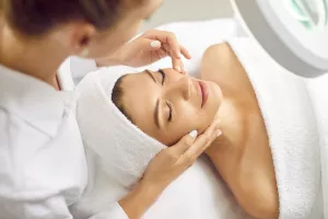 Young professional woman cosmetologist doing skin care facial treatment examining its condition for female client with special lamp in beauty clinic. Cosmetology and beauty procedure concept.